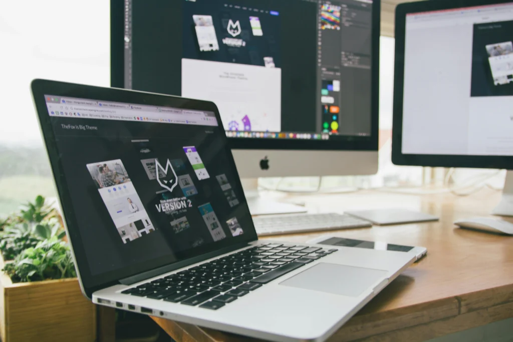 A laptop displays a website design for "Version 2," with two desktop monitors showing additional designs in a stylish workspace.